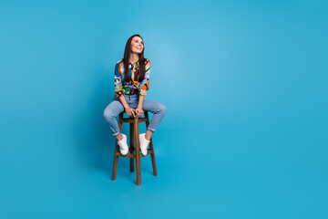 Full size photo of dreamy nice girl dressed print blouse jeans pants sit on chair look empty space isolated on blue color background
