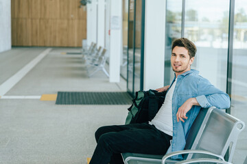 Young Caucasian male in modern transport hub sitting on bench with backpack. Casual attire with...