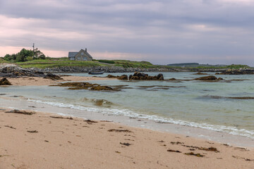 Gurteen Beach at sunset features a picturesque stone house overlooking a serene coastline in Ireland. The sandy shore, dotted with seaweed and rocks, meets the calm Atlantic waters