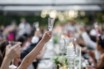 A raised champagne glass is held high in a celebratory toast during an outdoor event, with blurred attendees in the background and soft candlelight in the foreground