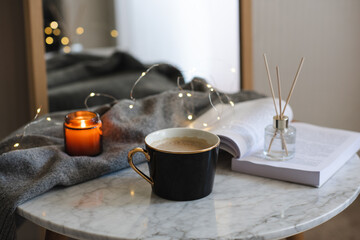 Cup of coffee with scented candle and open paper book on marble table over Christmas lights close up. Cozy home atmosphere.