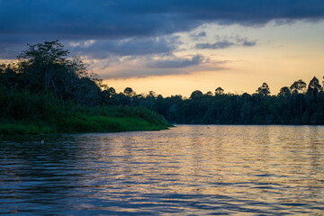 Light coming through dark stormy clouds behind riverbank of Kinabatangan River with blue light at dusk (Sunset), with river cruise boat in distance.
