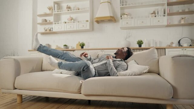 Carefree black man falling on soft couch in living room of modern apartment. Happy African American guy relaxing at home after hard working day, slow motion, cozy interior of house, relax in evening