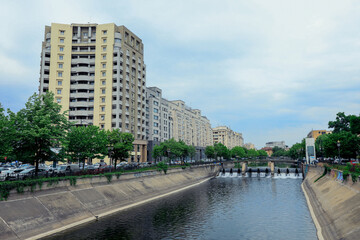 Fototapeta premium Waterway in Bucharest Romania lined with modern buildings and green trees during a cloudy day