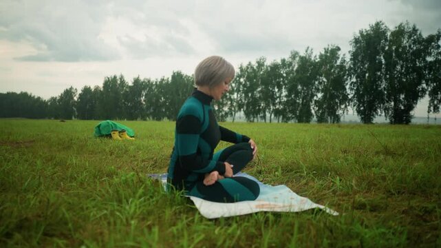 Side view of woman seated on yoga mat in siddhasana pose practicing yoga with yellow boots and green raincoat in the background, situated in a vast grassy field under cloudy sky lined with trees