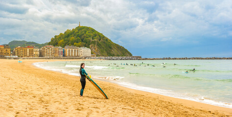 Fototapeta premium A surfer at Zurriola Beach with downtown San Sebastián and Monte Urgull in the background. Basque Country, Gipuzkoa, Spain.