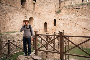 Retired man in hat, tourist traveler looking at ancient fortress as landmark making mobile phone call. Tourism and travel