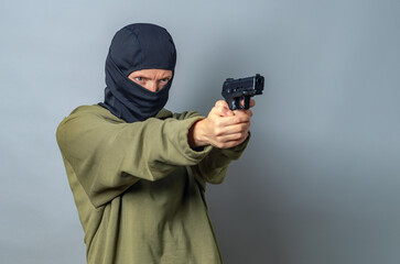 Woman in balaclava on head is holding gun in hands, aiming to right. Gray background. Copy space.