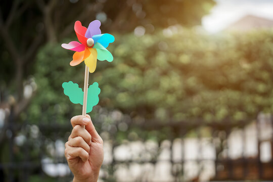 A woman joyfully holds a colorful pinwheel and a flower in her hands while standing in a vibrant garden under a blue sky on a breezy summer day