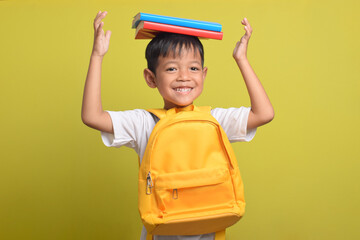 Back to school. Cheerful Asian boy with backpack and carrying books on his head isolated on yellow...