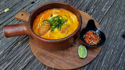 Laksa, spicy noodles served in a pot with lime wedges and sambal. Isolated wooden background.