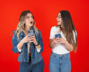 Looking at each other, portrait of young caucasian two women looking at each other and using mobile cell phone shocked face expression over red studio background copy space. 