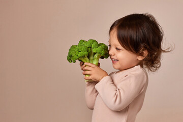 smiling little child girl holding fresh green raw broccoli on beige background. healthy baby food....