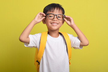 Portrait of Asian boy smiling with yellow backpack, posing holding his glasses, isolated on yellow background. Looking at camera. Education concept. Copy space