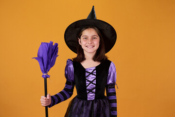 Smiling youthful girl with purple broom standing in isolation against orange background and looking...