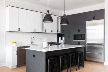 A modern kitchen with white and black cabinets, bar stools sitting at a large island, black pendant lights hanging over the marble countertop, and a subway tile backsplash. No brands or labels.