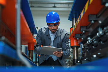 African American engineer with a blue helmet holding a tablet, doing factory working machine...
