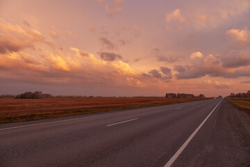 Road Leading Into A Sunset. View of Country road passing through fields during sunset. Asphalt road in rural landscape at sunset with dramatic clouds.