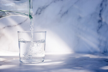 Pouring drinking water into glass on white table in morning sunlight.