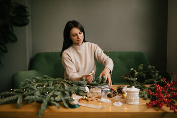 Woman making Christmas wreath using natural pine branches, sitting near wooden table.