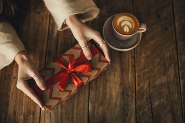 Woman holding Christmas present wrapped in brown paper with ribbon, drinking cappuccino by wooden table. Hands close up.