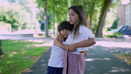 Fototapeta premium Boy hugging the girl in a warm embrace on a park pathway. The outdoor setting and their expressions suggest a moment of comfort and sibling bonding