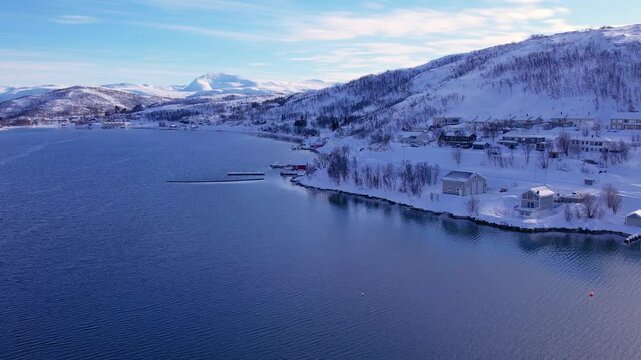 Snow covered mountain range on coastline in winter, Norway. Surroundings of town Tromso. Panoramic aerial view landscape of nordic snow cowered mountains, houses and ocean. Troms county, Fjordgard