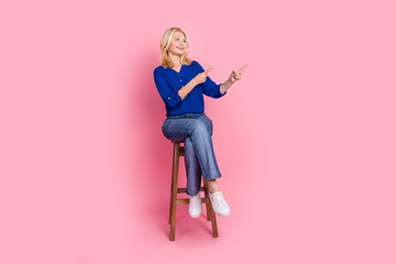 Fototapeta premium Full length photo of lovely pensioner lady sit chair look point empty space dressed stylish blue garment isolated on pink color background
