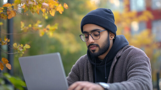 A man wearing a black hat and glasses is sitting in front of a laptop