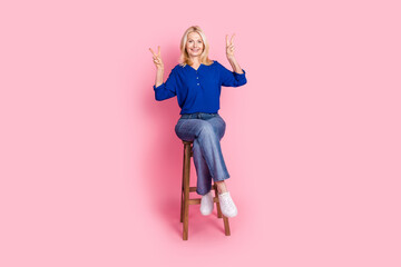 Full length photo of lovely pensioner lady sit chair show v-sign dressed stylish blue garment isolated on pink color background