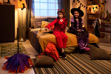 Two youthful children in Halloween costumes sitting on bed and looking at moving broom on vacuum cleaner being controlled by girl