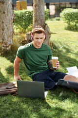 Obraz premium A young man with a prosthetic leg sits on the grass, sipping coffee while working on his laptop.