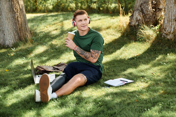A young man with a prosthetic leg relaxes in a park, working on his laptop and sipping a drink.