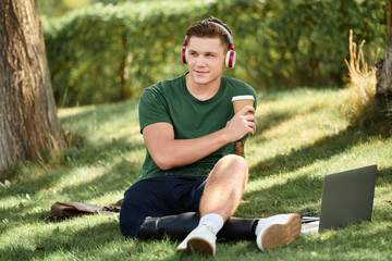 A young man relaxes on the grass, sipping coffee while working on his laptop, embodying inclusion...