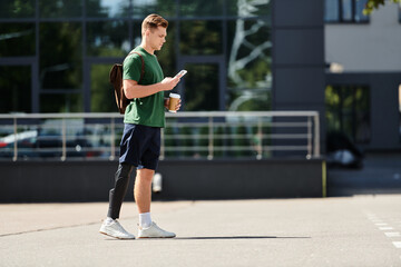 Obraz premium A young man with a prosthetic leg stands outdoors, sipping coffee and focused on his phone in the sunlight.