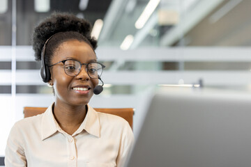 Confident African American businesswoman wearing headset working on laptop in modern office. Engaged in remote communication, customer service, or virtual meeting. Professional demeanor displayed.