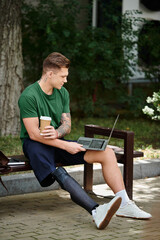 Obraz premium A young man with a prosthetic leg relaxes on a bench, sipping coffee and focused on his laptop in a park.