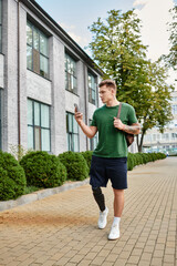 A young man with a prosthetic leg strolls along a sunny pathway, engaging with his smartphone.