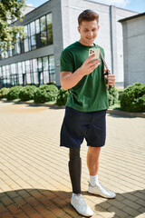 A young man with a prosthetic leg smiles while looking at his phone in an outdoor setting filled...