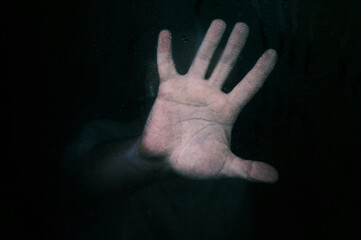 Horror scene of a man with hand against wet glass on dark background.