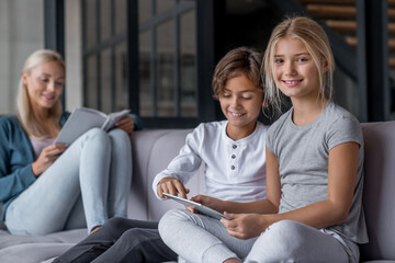 Children using digital tablet watching video cartoons playing games while mother reading book sitting resting on sofa in living room. Babysitter nanny tutor teacher spending time with kids at home.