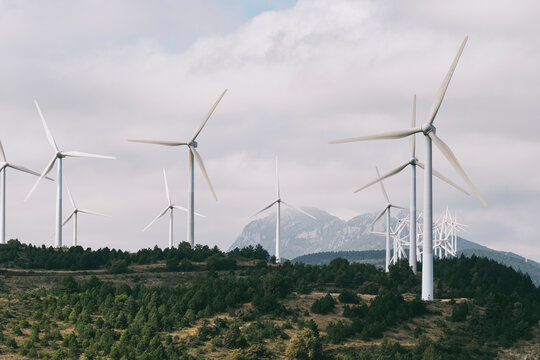 A cluster of towering wind turbines stands firm at the base of a mountain range on a partially cloudy day, representing clean energy and modern sustainability solutions in Navarra Spain