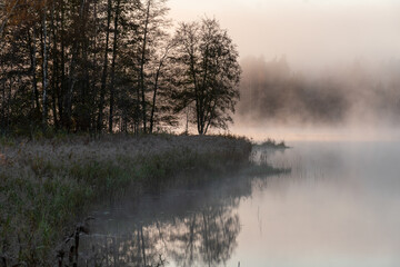 Blurred landscape with a lake in the morning fog, fog over the lake, dark silhouettes of trees on the shore of the lake