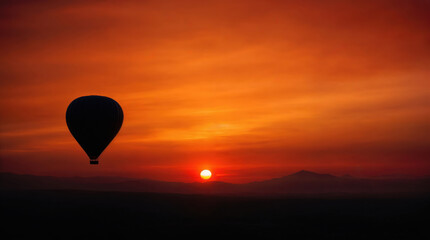 Fototapeta premium Silhouette of hot air balloon floating against dramatic orange sunset sky and mountain landscape, atmospheric adventure travel scene