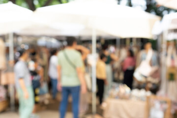 A group of people are standing outside under a white umbrella. The scene is blurry, giving it a sense of movement and energy. The people are browsing through a market, with various items on display