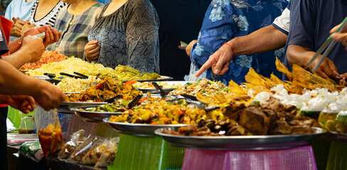 A group of people are gathered around a buffet table with a variety of food. Scene is lively and...