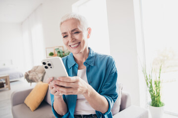 Photo of nice aged lady toothy smile use smart phone wear blue shirt bright interior apartment indoors