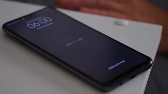 Man waking up, turning off alarm clock to snooze mode on smartphone screen. Sleepy man touching phone with alarm at 6 o'clock morning