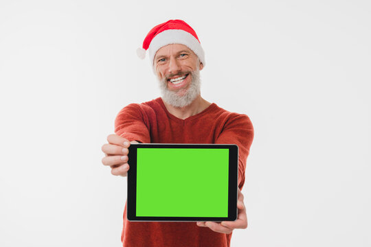 Happy mature middle-aged man in red Santa Claus hat holding digital tablet showing green screen for mockup copy space isolated on white background