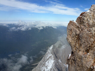 A view of distant mountains below and white clouds above them in a blue sky. To the right is a stony slope of a rocky mountain